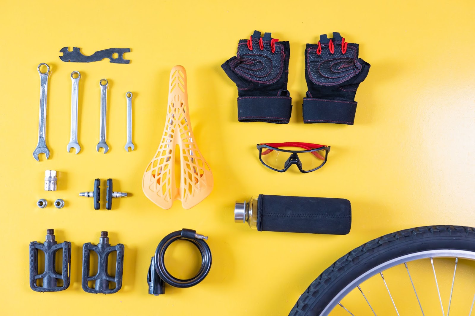 A flat lay of various bike accessories and tools on a yellow background, including a mountain bike saddle, pedals, wrenches, gloves, and a cable lock, suitable for maintaining both mountain bike vs. racing bike setups. The arrangement showcases the different tools used for each bike type.