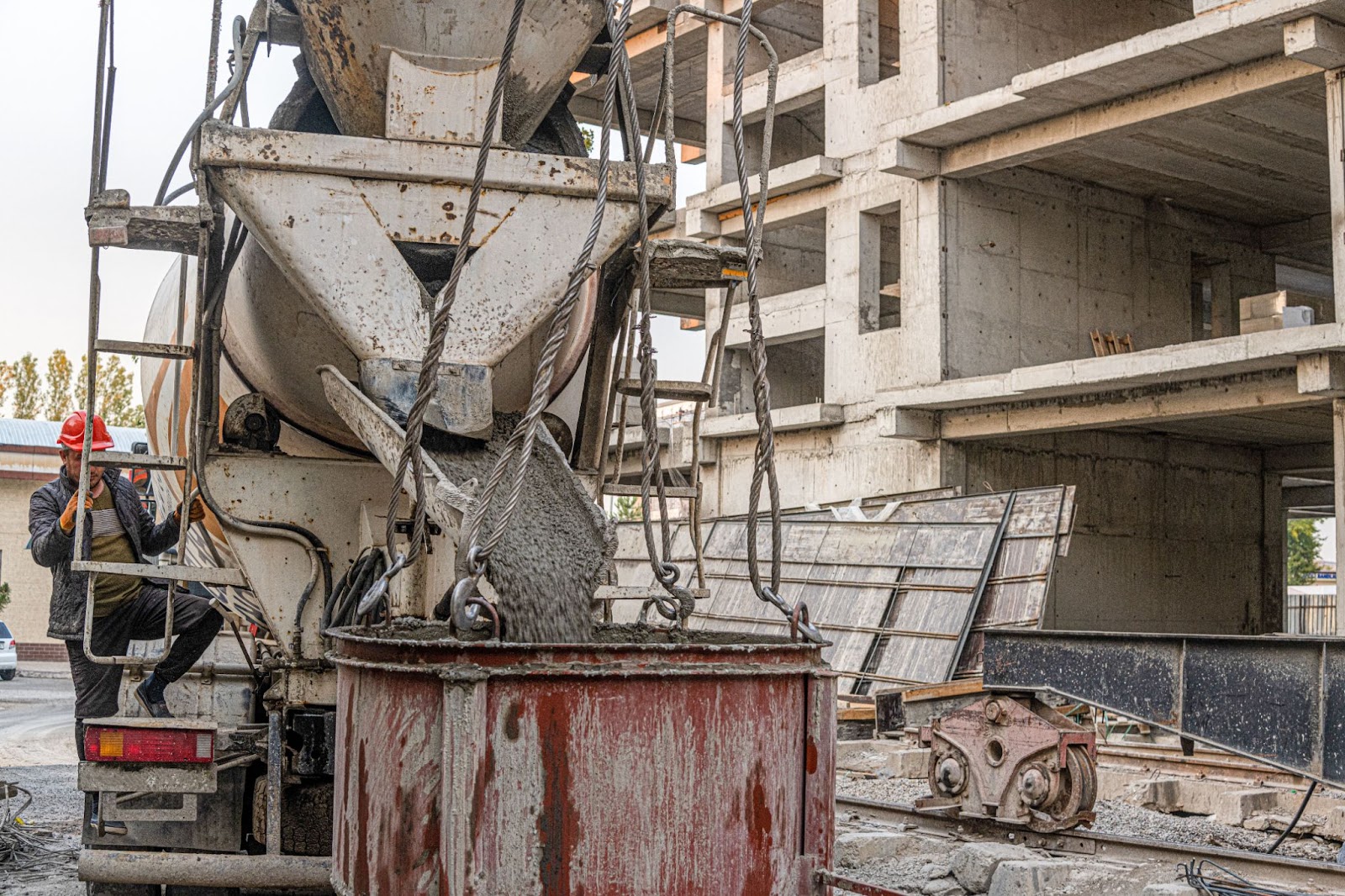 A worker operates a concrete pouring bucket at an industrial construction site, with the building structure visible in the background.