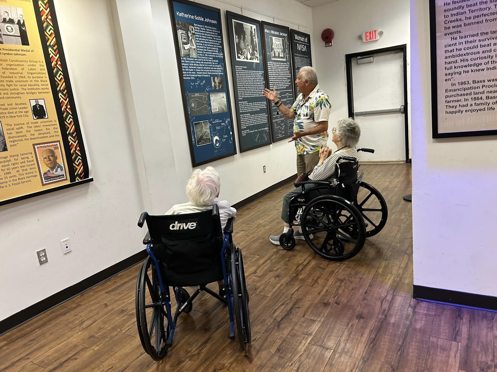 Two people in wheelchairs at a museum with a guide explaining the exhibits