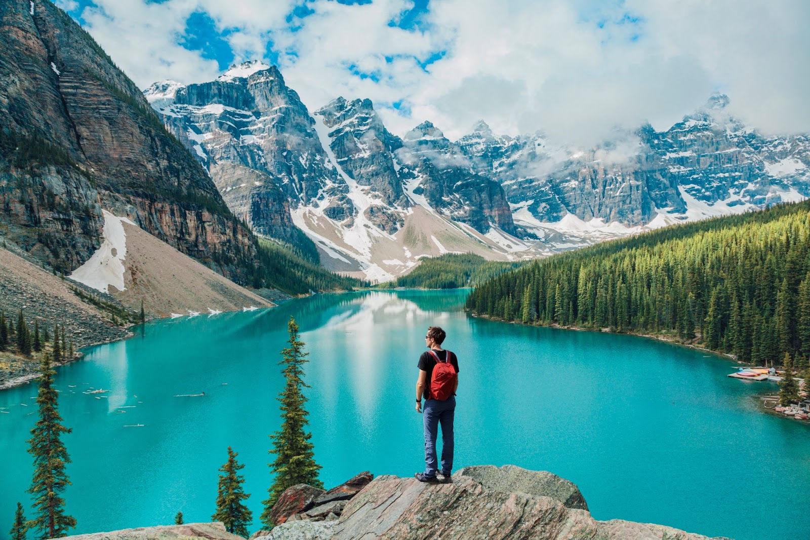 A man with a red backpack stands on a rock overlooking a turquoise lake surrounded by snowy mountains and evergreen forests at Glacier National Park