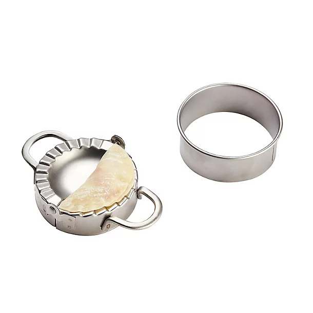 A stainless steel dumpling press with a half-sealed dumpling inside, alongside a matching round dough cutter.