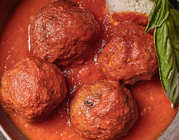 Four large meatballs covered in rich tomato sauce, garnished with fresh basil, and served with a side of bread in a deep dish.