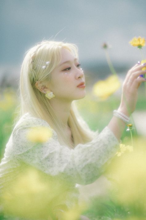 This contains an image of Maknae Haeun's with a long blonde hair holding a flower in her hand and looking up at the sky