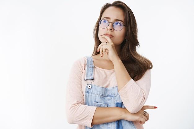 Garota fazendo suposição resolvendo mistério em mente segurando o dedo no lábio olhando para o canto superior esquerdo determinado e pensativo como pensando tomando decisão ou escolha em pé focado sobre fundo cinza