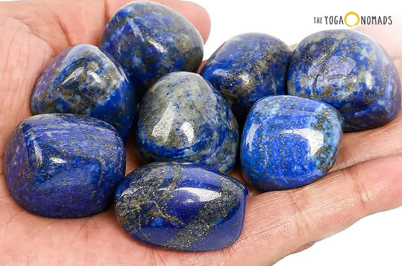 A collection of polished lapis lazuli stones being held in a person’s hand. These stones are primarily deep blue in color with flecks of golden pyrite and white calcite throughout. The stones are smooth and have an oval shape, varying slightly in size and pattern.