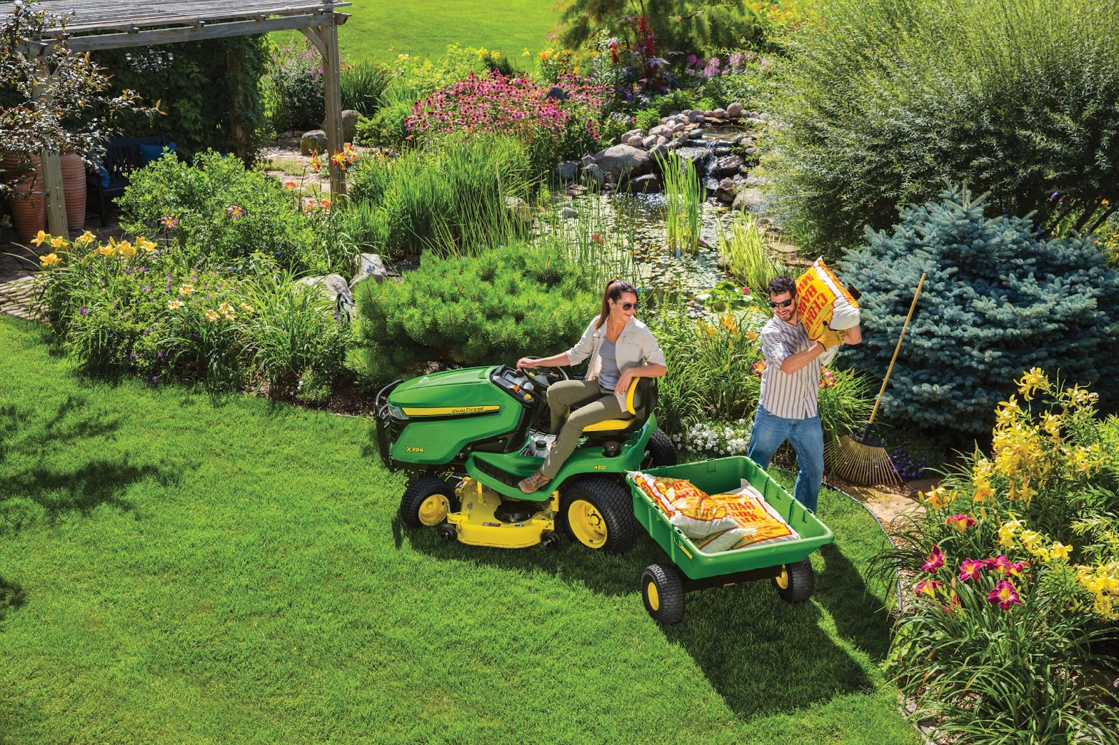 A couple enjoying yard work in their vibrant garden, using a John Deere lawn tractor towing a utility cart filled with gardening supplies, showcasing efficiency and ease of yard maintenance.