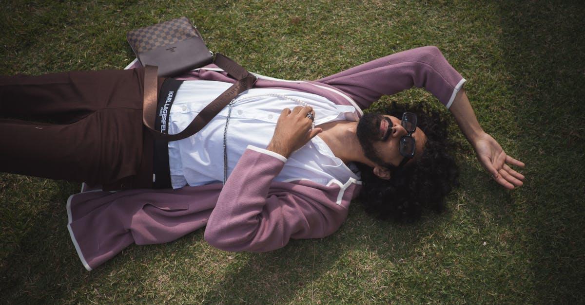 A fashionable man with curly hair lies on grass wearing sunglasses and a purple blazer.