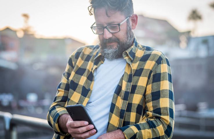 Mature bearded man use mobile phone with internet wireless connection wearing glasses. Adult male people chatting with smartphone outside. Sunlight in background