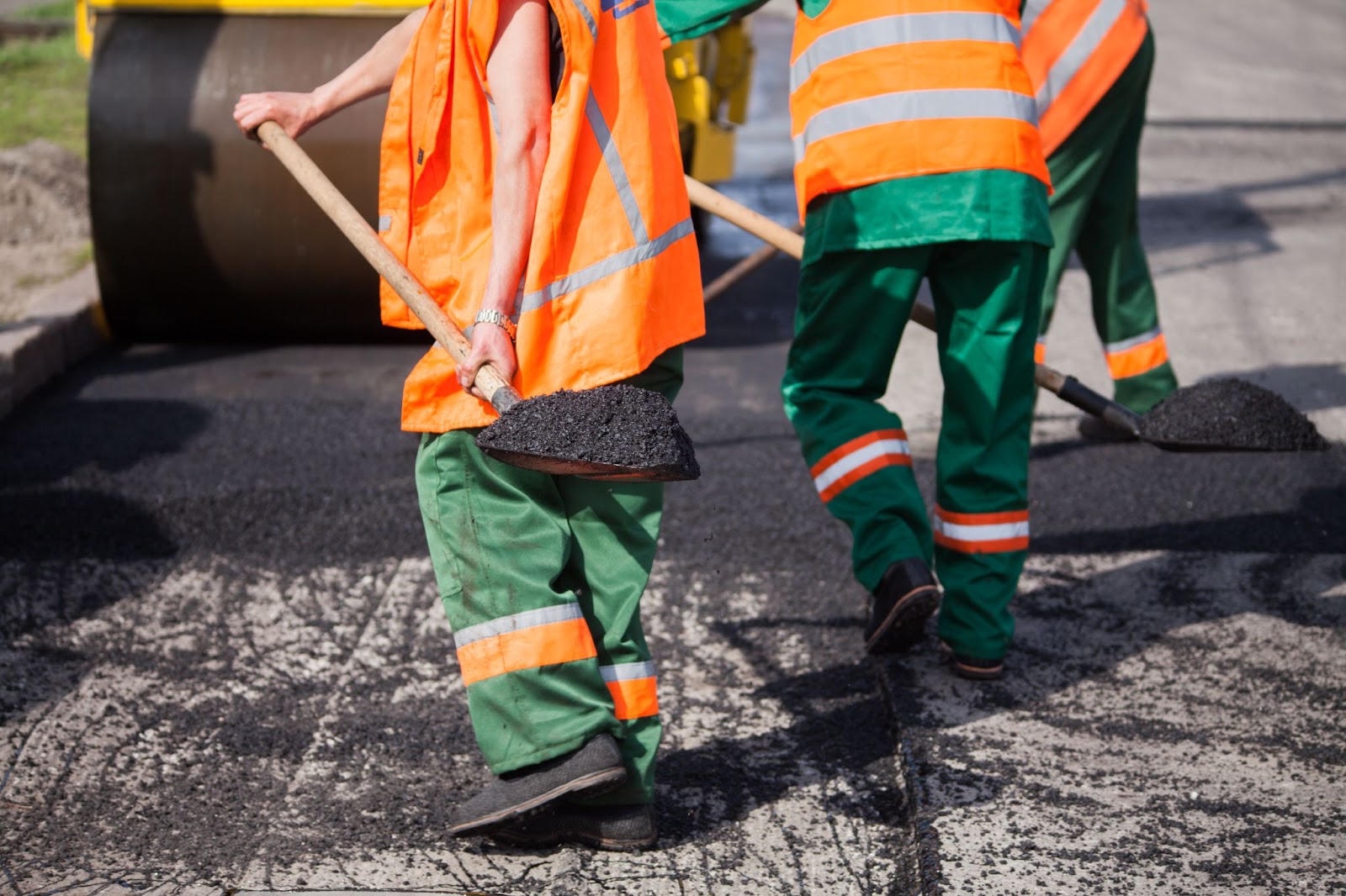 Three workers in orange, green, and white safety gear patch potholes with asphalt on a roadway.