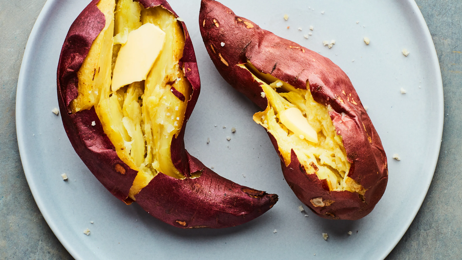 Two roasted sweet potatoes split open and topped with melting butter, sprinkled with sea salt, served on a gray plate.