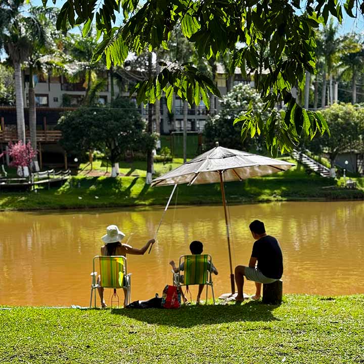Família desfrutando de um dia ao ar livre na orla de um rio, sob guarda-sol, com árvores ao redor e ambiente tranquilo na natureza brasileira.