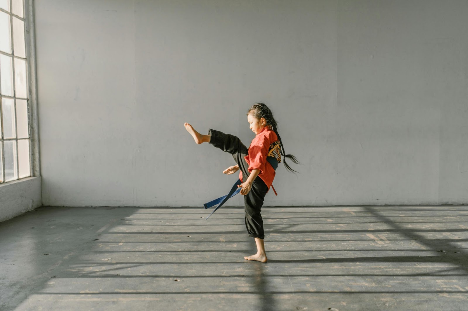 A young martial arts student performing a high kick with her left leg, wearing a traditional martial arts uniform in a training studio