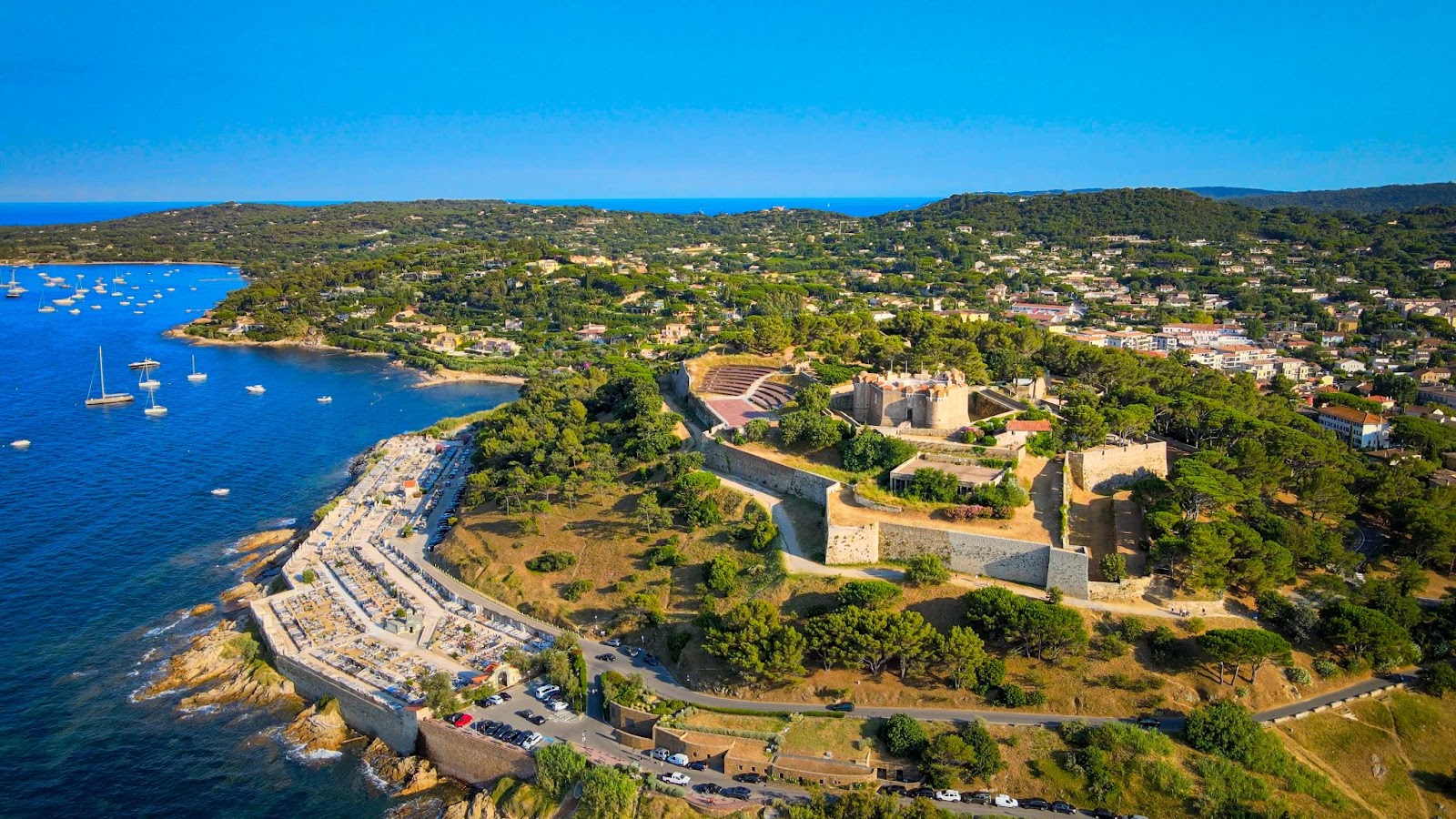 Aerial view of the Citadel of Saint-Tropez, a coastal fortress surrounded by lush greenery, overlooking the sea with anchored boats and the town of Saint-Tropez in the background.