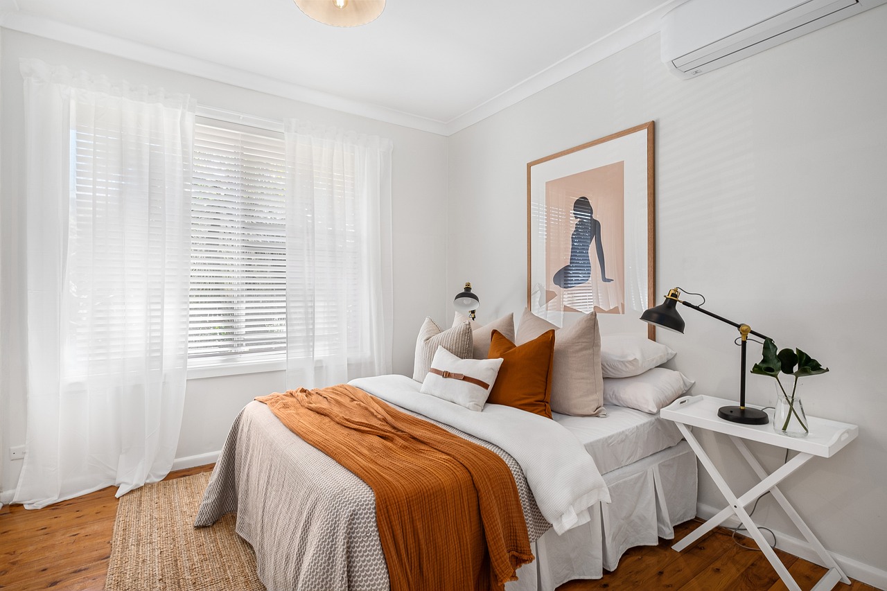 Bedroom with white and orange bedding, a black lamp on a white nightstand, white curtains, and a silhouette artwork on the wall.