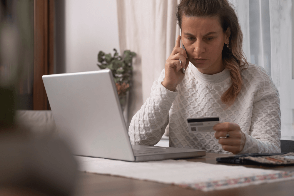 mulher de blusa branca preocupada com cartão na mão e falando ao telefone após ter sofrido golpe virtual