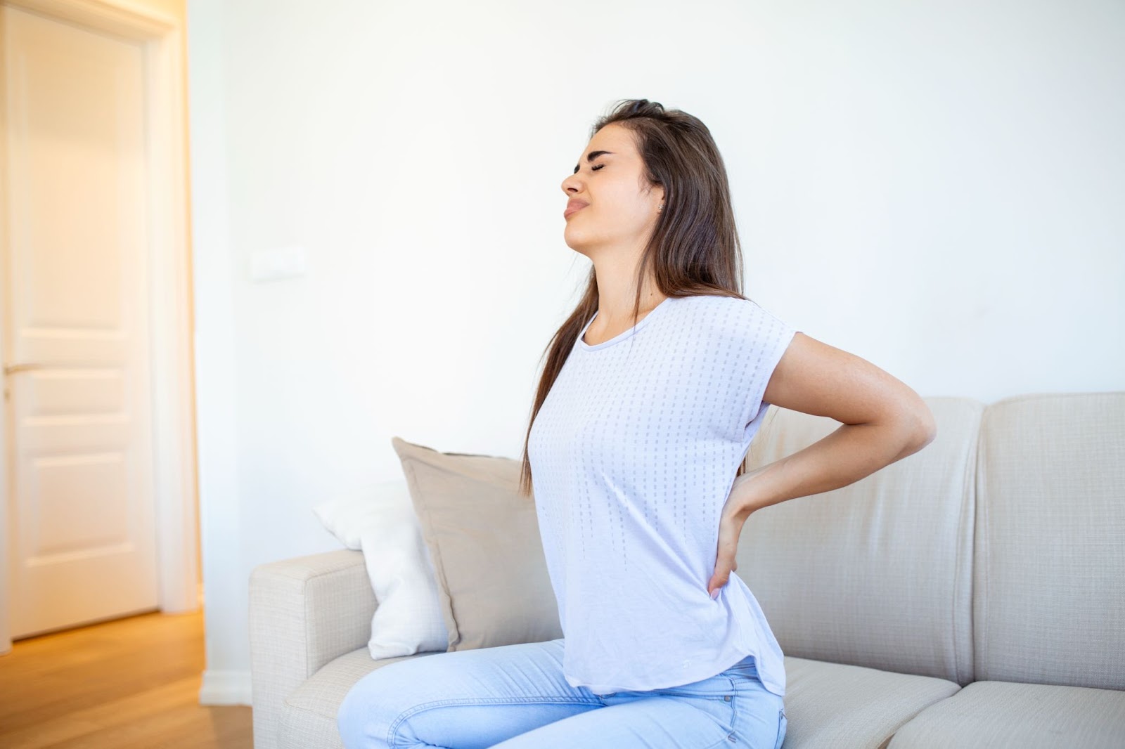 A woman sitting on a couch holds her back in pain.