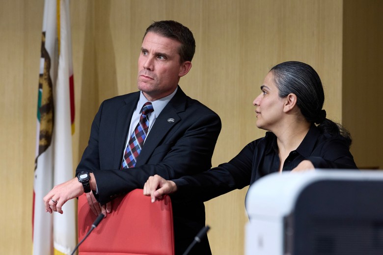 Two individuals stand near a podium in a formal setting. One is leaning on the back of a red chair, looking thoughtfully into the distance, while the other gestures toward them in conversation. A California state flag is partially visible in the background.