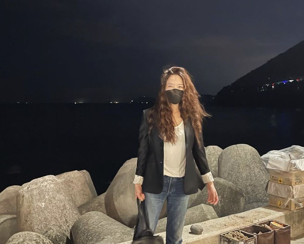 Former Miss Korea Kim Min Kyung  wearing a black jacket and a white shirt and a blue jean trouser putting on a nose marks standing in front of rocks 