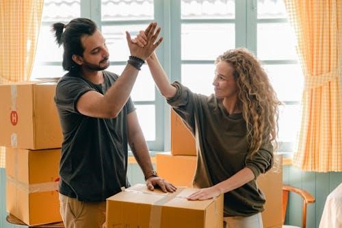 Free Happy woman in casual wear standing near heap of cardboard boxes and giving high five to ethnic boyfriend with ponytail showing agreement while looking at each other Stock Photo