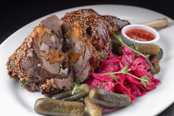 A plate featuring a roasted lamb shank crusted with mustard seeds, served with pickles, vibrant red cabbage, and a side of dipping sauce