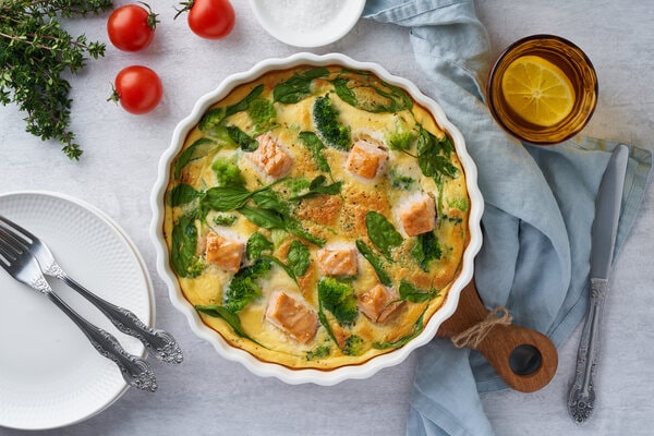 Smoked salmon frittata served in a white dish alongside utensils, cherry tomatoes, and a refreshing drink, set on a light tablecloth