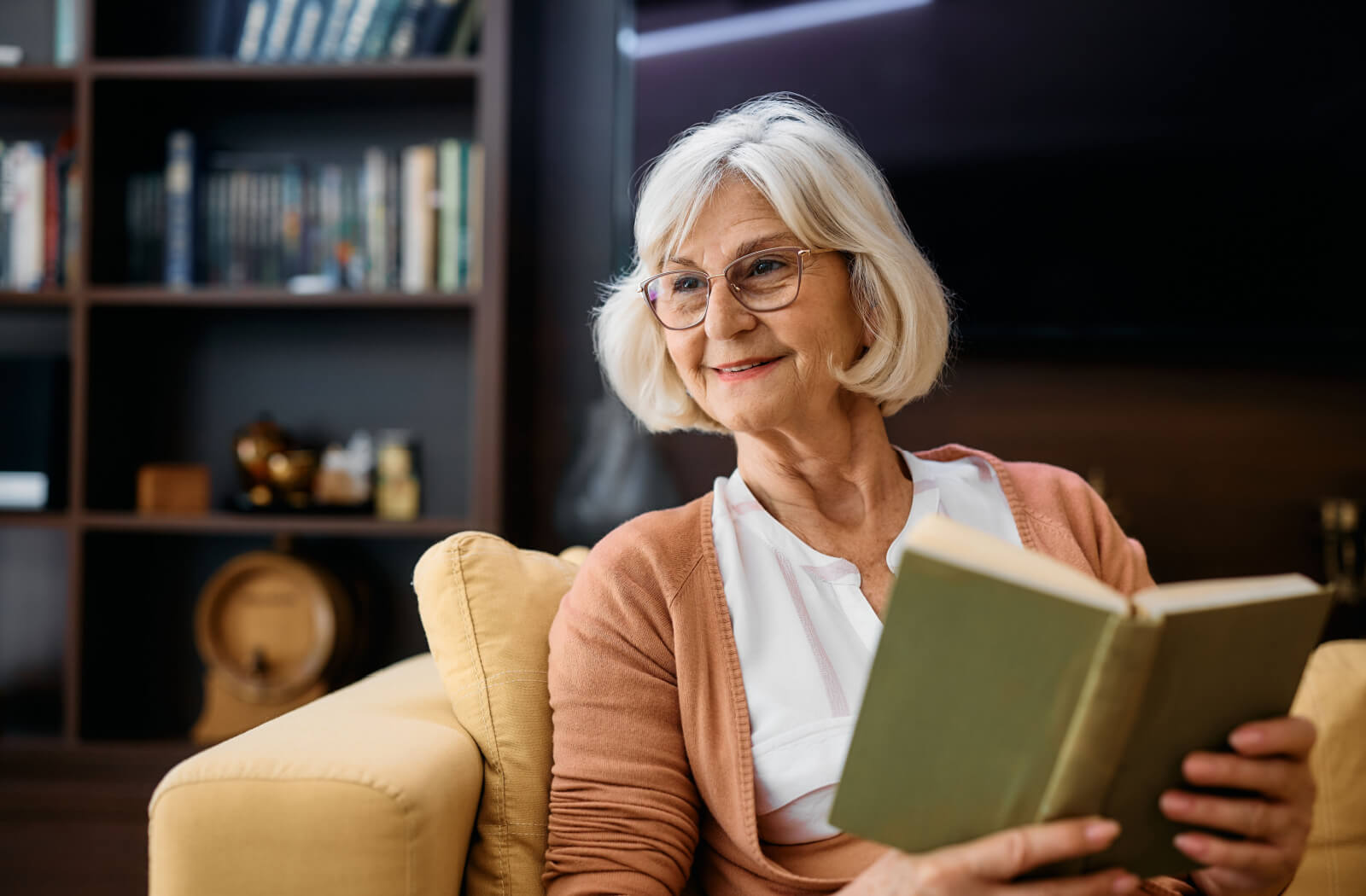 An older adult smiling while sitting on the couch at home reading a small green book.