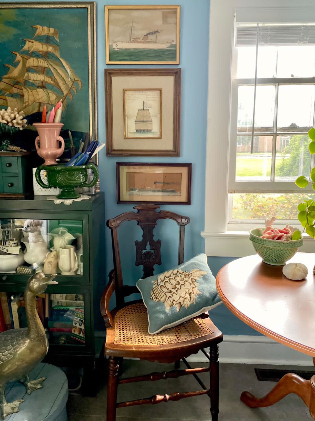 Couple of ship paintings hanging next to a window and above a green cabinet which is placed next to an armchair and is filled with lots of white porcelains and books. On top of the cabinet is an assortment of decorative items.