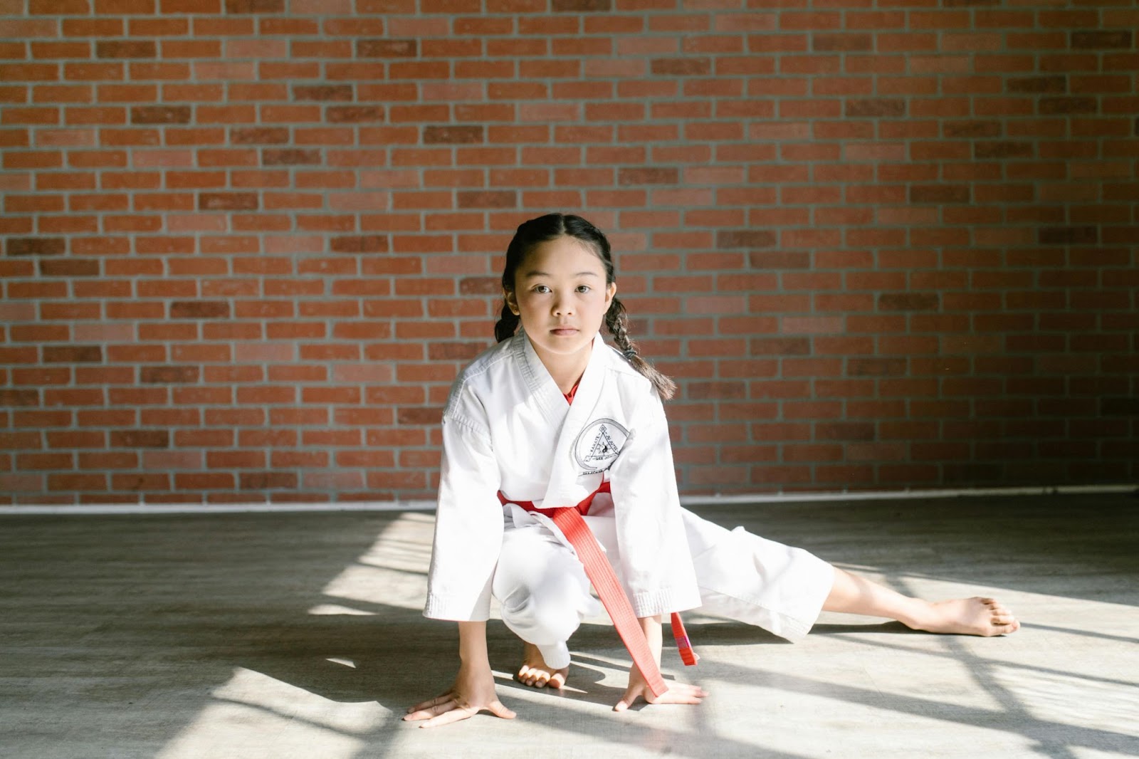 A young girl in a martial arts uniform holds a low stance, with both hands on the ground and one leg stretched out to the side, showing balance and flexibility.