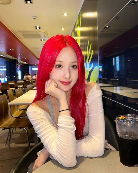 This  contain Belle, a member of the K-pop group KISS OF LIFE, with long red hair sitting at a table in a restaurant next to a drink