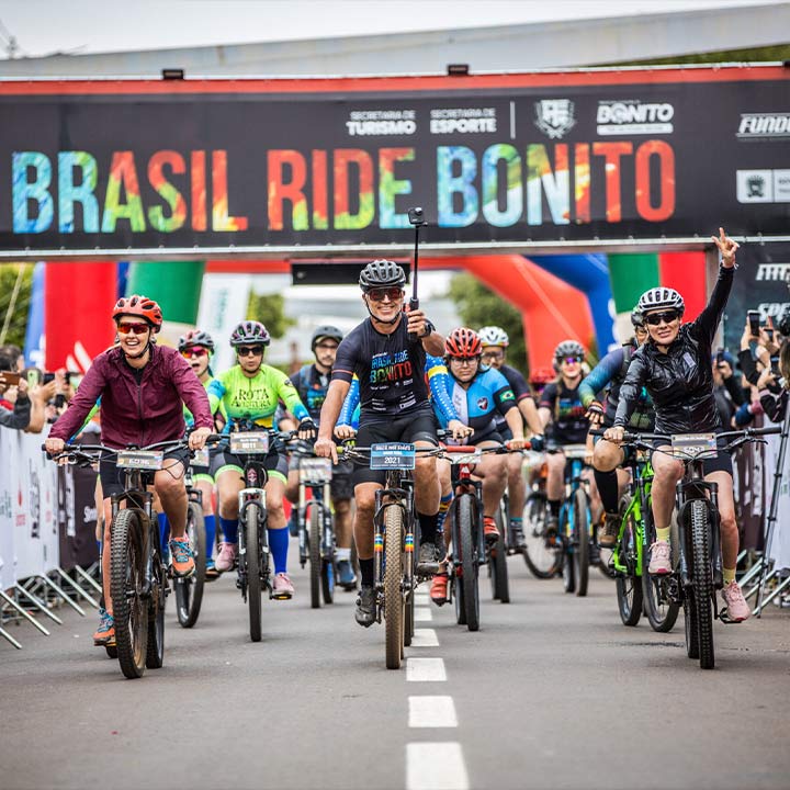 Grupo de ciclistas comemorando a chegada na linha de chegada no evento Brasil Ride Bonito, com diversas pessoas usando capacete e vestimentas esportivas, sob um arco de chegada colorido que celebra o turismo de aventura e o esporte no Brasil.