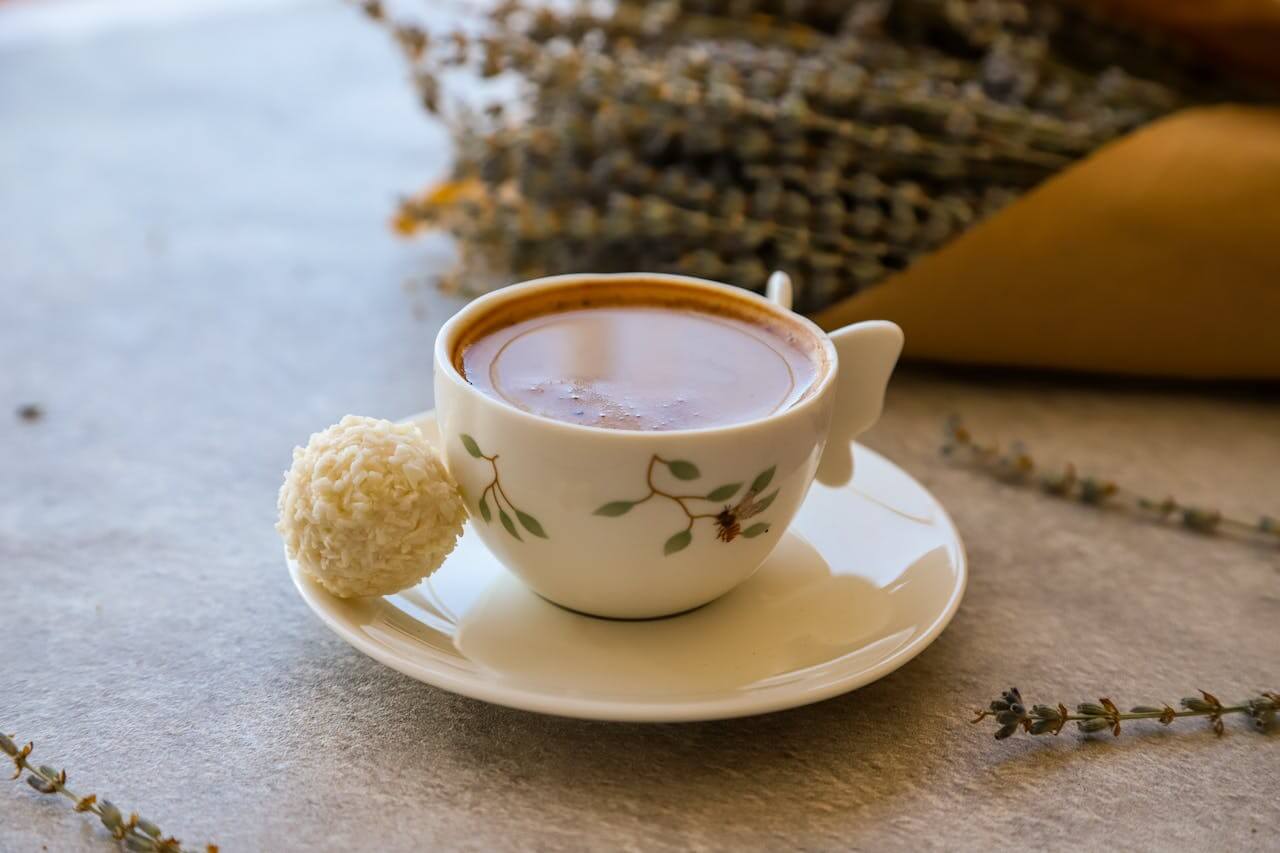 A small, ornate porcelain cup filled with dark coffee sits on a white saucer. A single white chocolate truffle rests beside the cup. A bouquet of dried lavender is blurred in the background. The image evokes a sense of calm and relaxation, perfect for a cozy morning or afternoon.