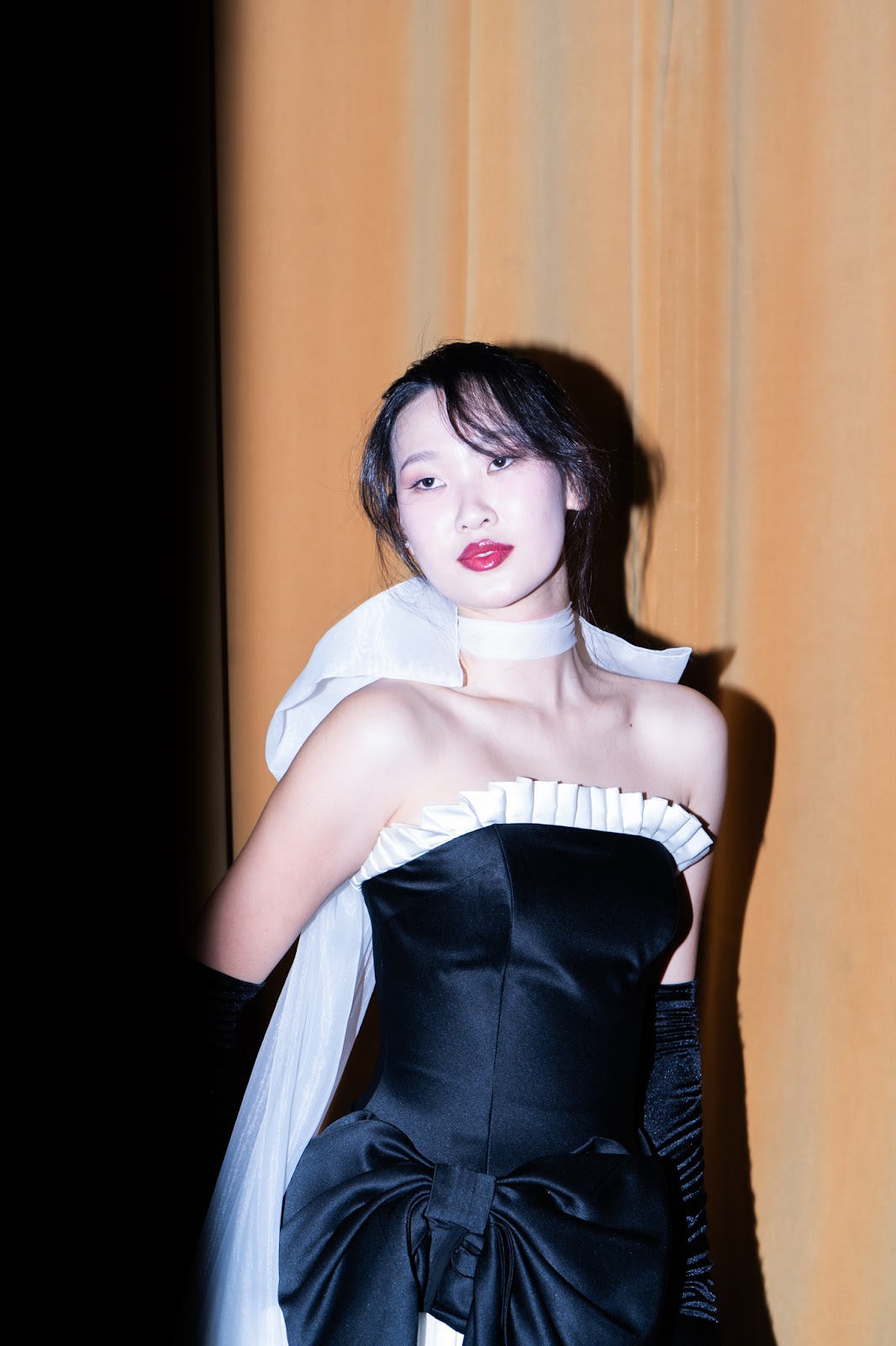 Enkhjin Batbold illuminated against a warm golden curtain and looking off to the side, showing the details of a black-and-white gown and sheer bow collar. 