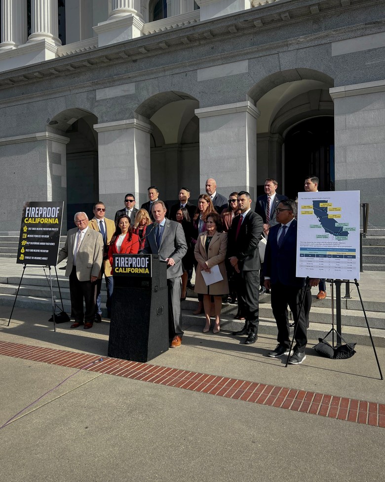 A group of individuals stands on the steps of a government building during a press conference. A speaker addresses the audience from a podium with a sign reading 'Fireproof California.' Two large posters with text and maps flank the group. The building's arched entrances and columns serve as the backdrop.