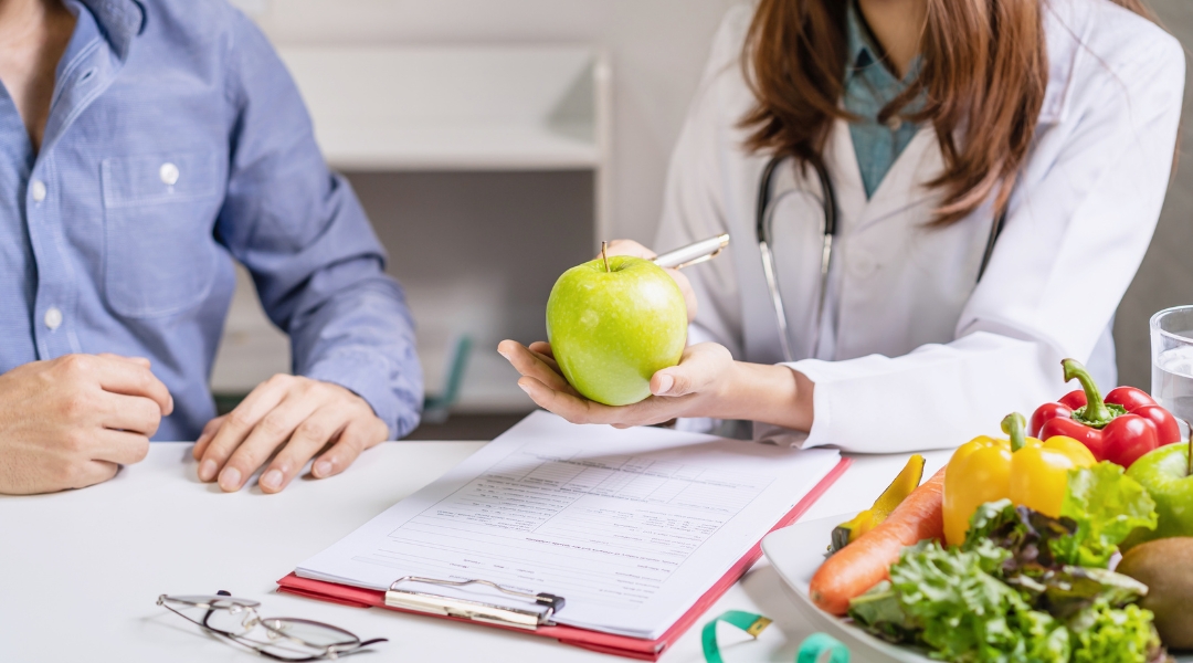 Nutritionniste vêtue d'une blouse blanche, tient une pomme verte tout en expliquant quelque chose à un patient. Sur la table, des documents et un plateau de fruits et légumes frais, notamment des carottes, des poivrons et de la salade, sont disposés.