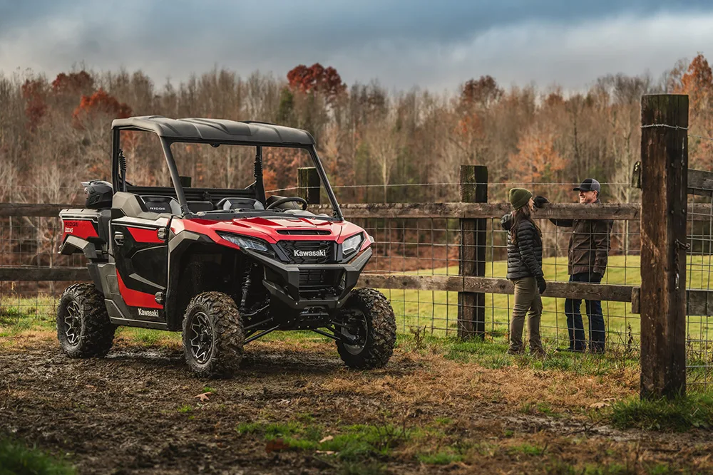 A 2025 Kawasaki Ridge parked on grass while its driver speaks to a friend over a fence nearby.