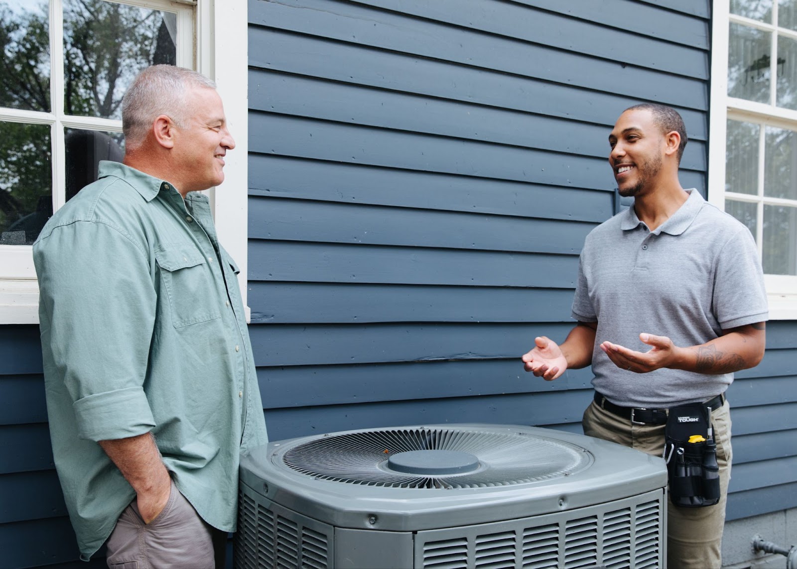 Two men discussing heat pumps and energy efficiency upgrades, standing outside of a dusty blue home with a heat pump.