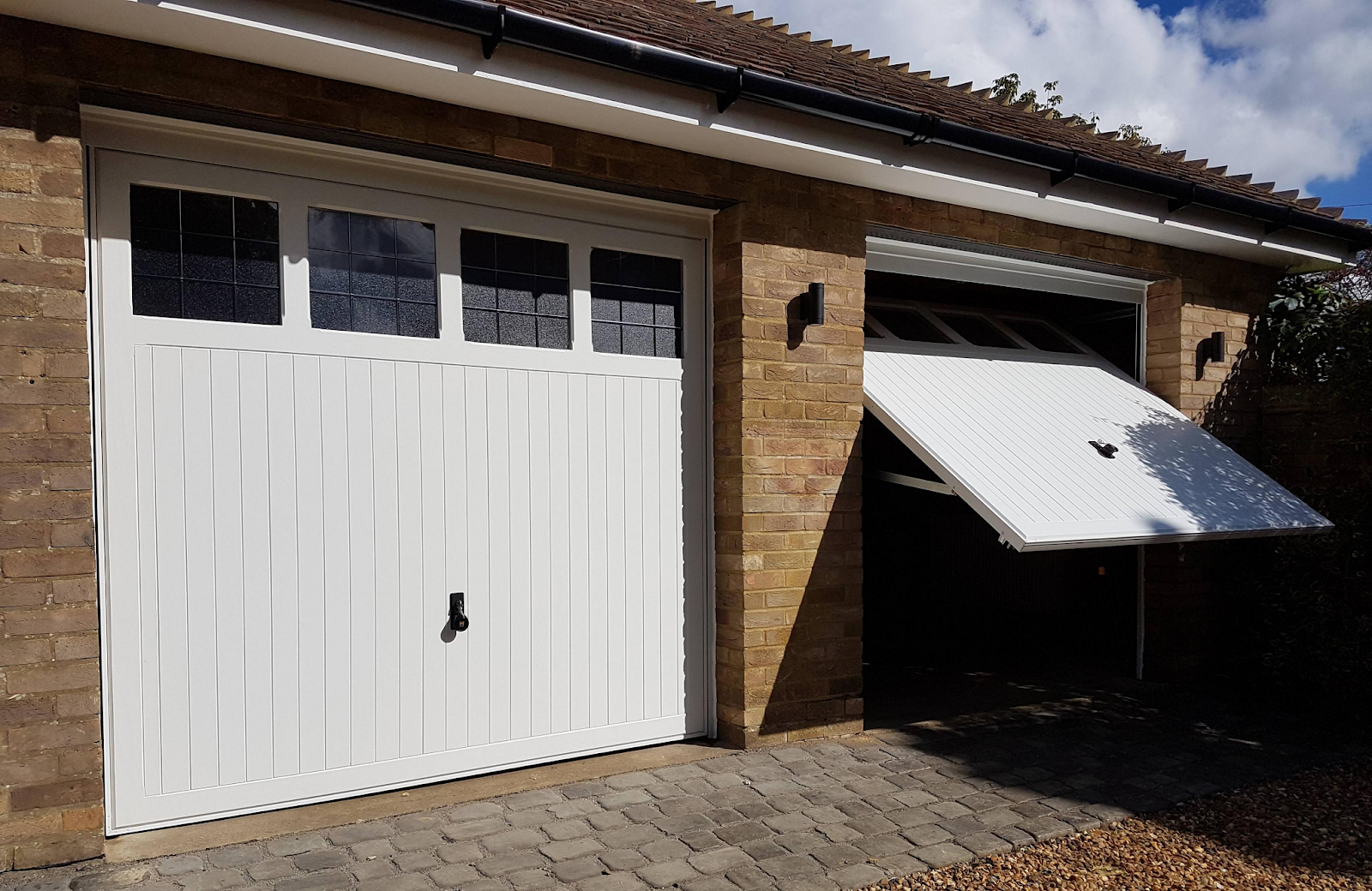 A brick garage with two white up-and-over garage doors, one closed and the other partially open, featuring decorative glass panels at the top, set against a paved driveway on a sunny day