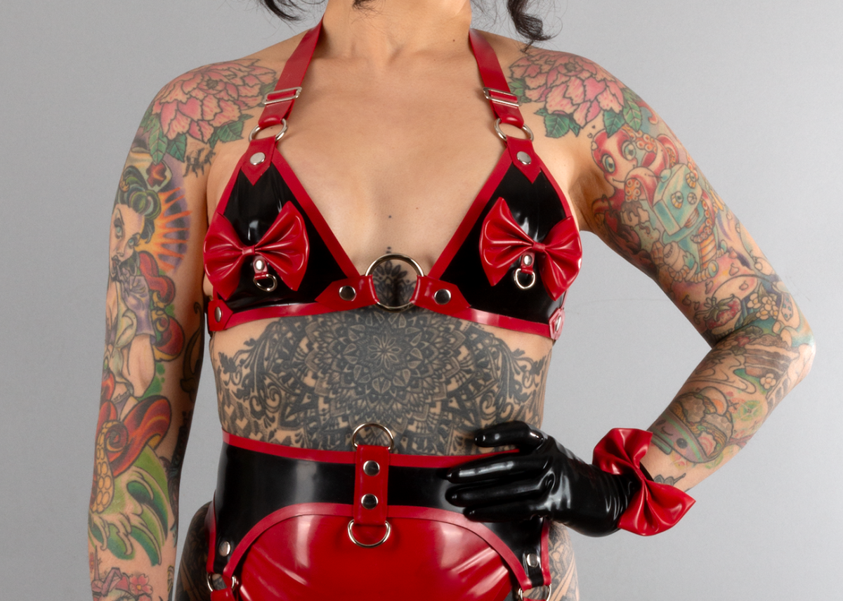 A tattooed femme models a black and red lingerie set with matching black gloves, all made of latex