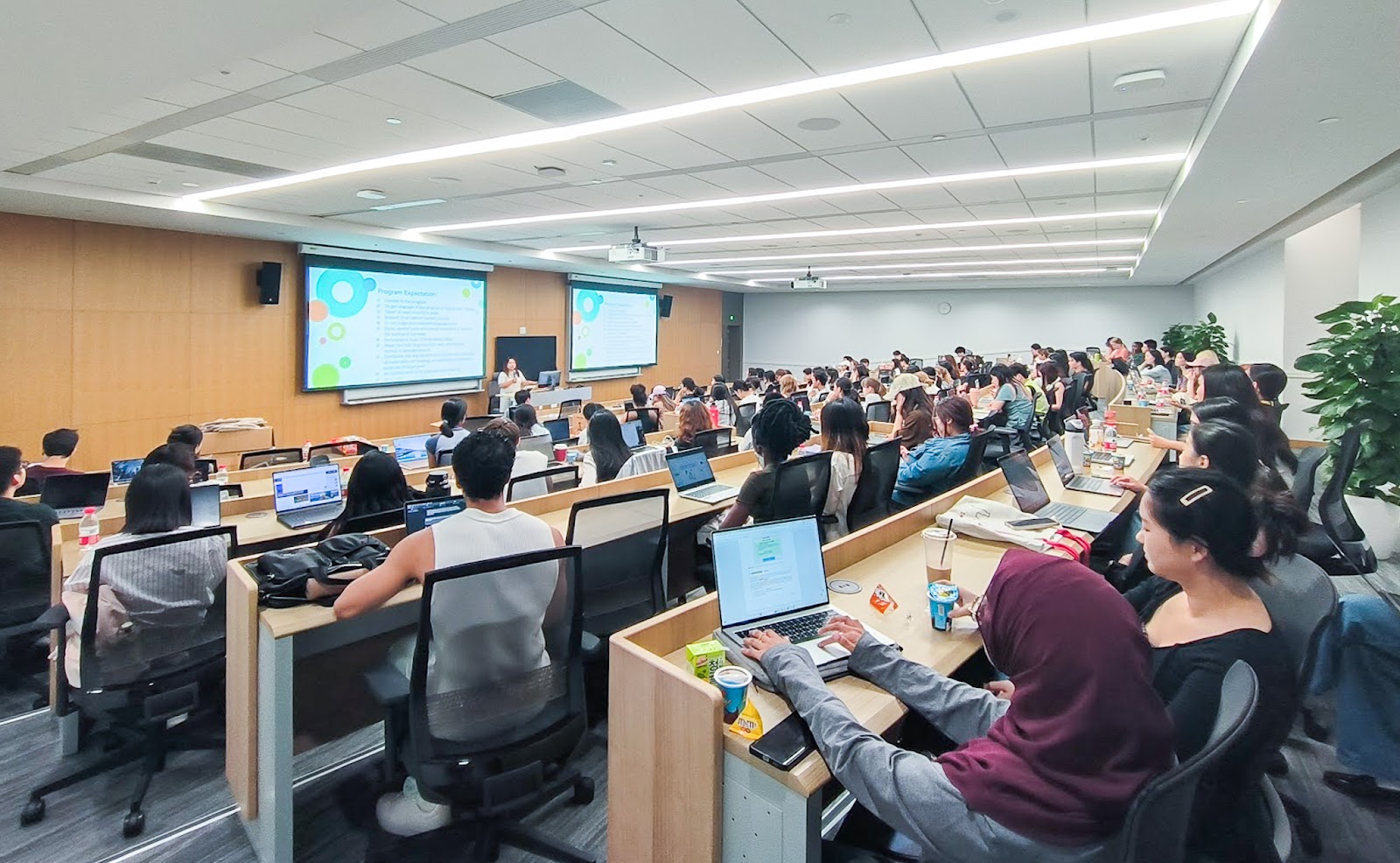 rows of students sitting in a classroom facing a speaker and projector screens at the front of the room