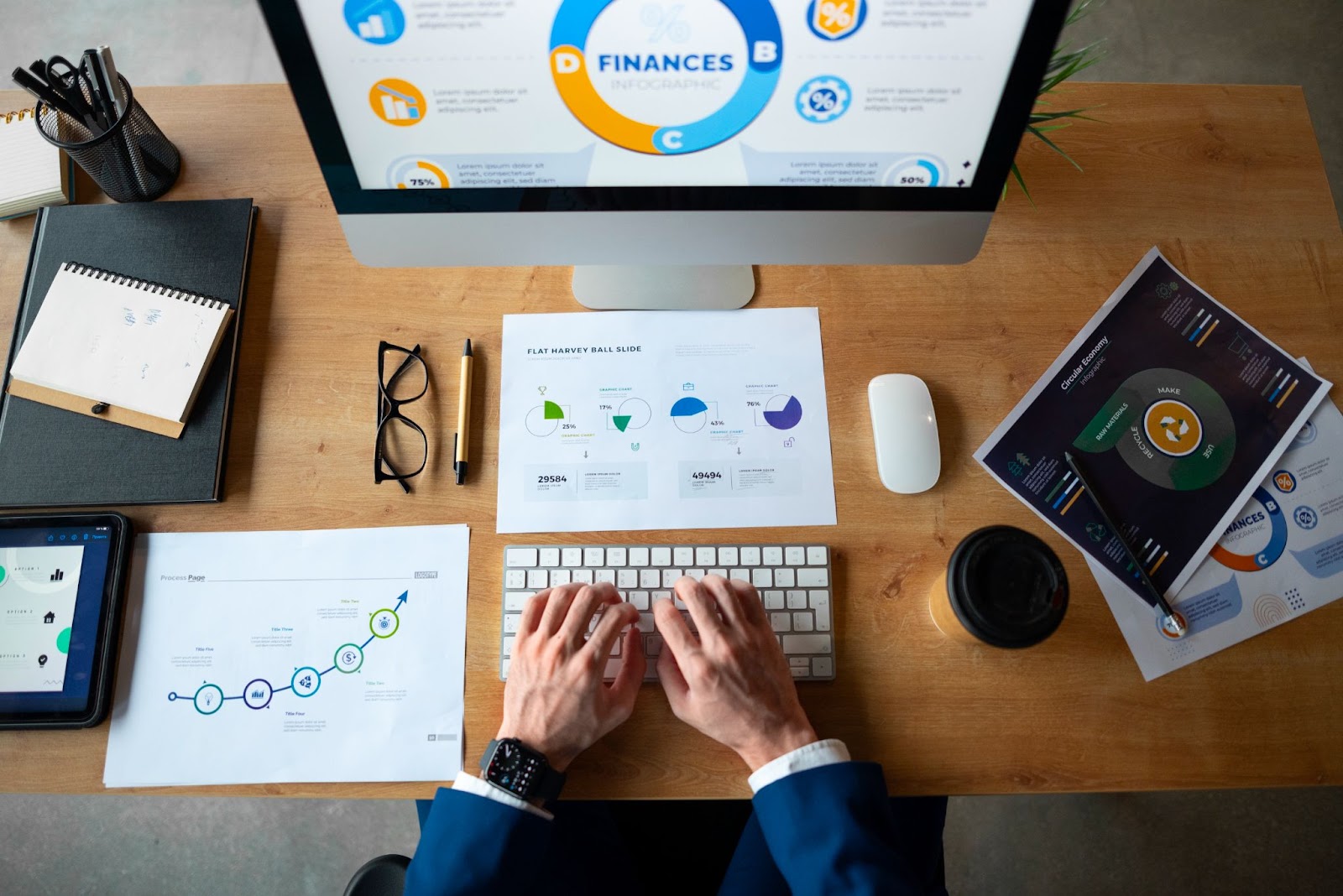 Top view of a workspace with a desktop monitor displaying finance infographics, printed analytics reports, and hands typing on a keyboard representing digital financial planning and data-driven decision-making.