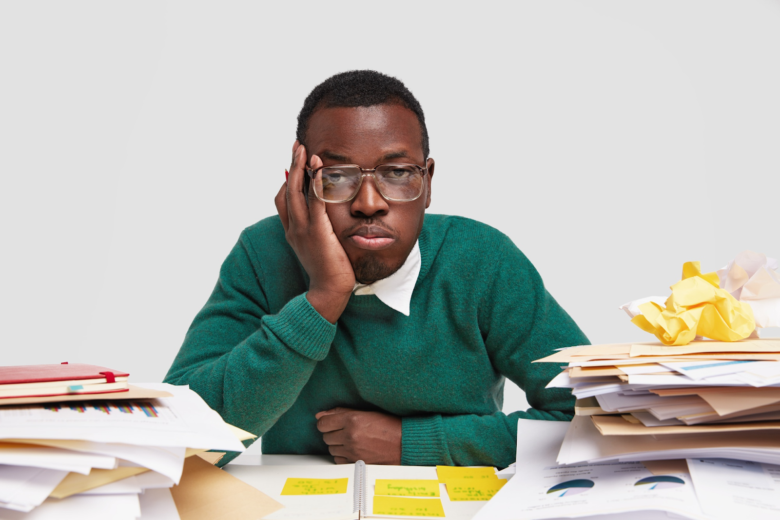 A man seated amongst a pile of papers on his desk with his hand on his chin contemplating what steps to take. 