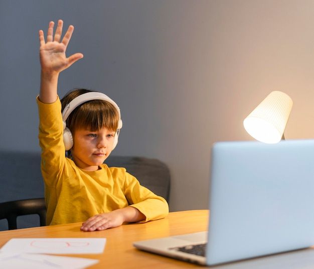 Young boy in a yellow shirt attending an online class, raising his hand, wearing headphones, and sitting at a desk with a laptop and a table lamp