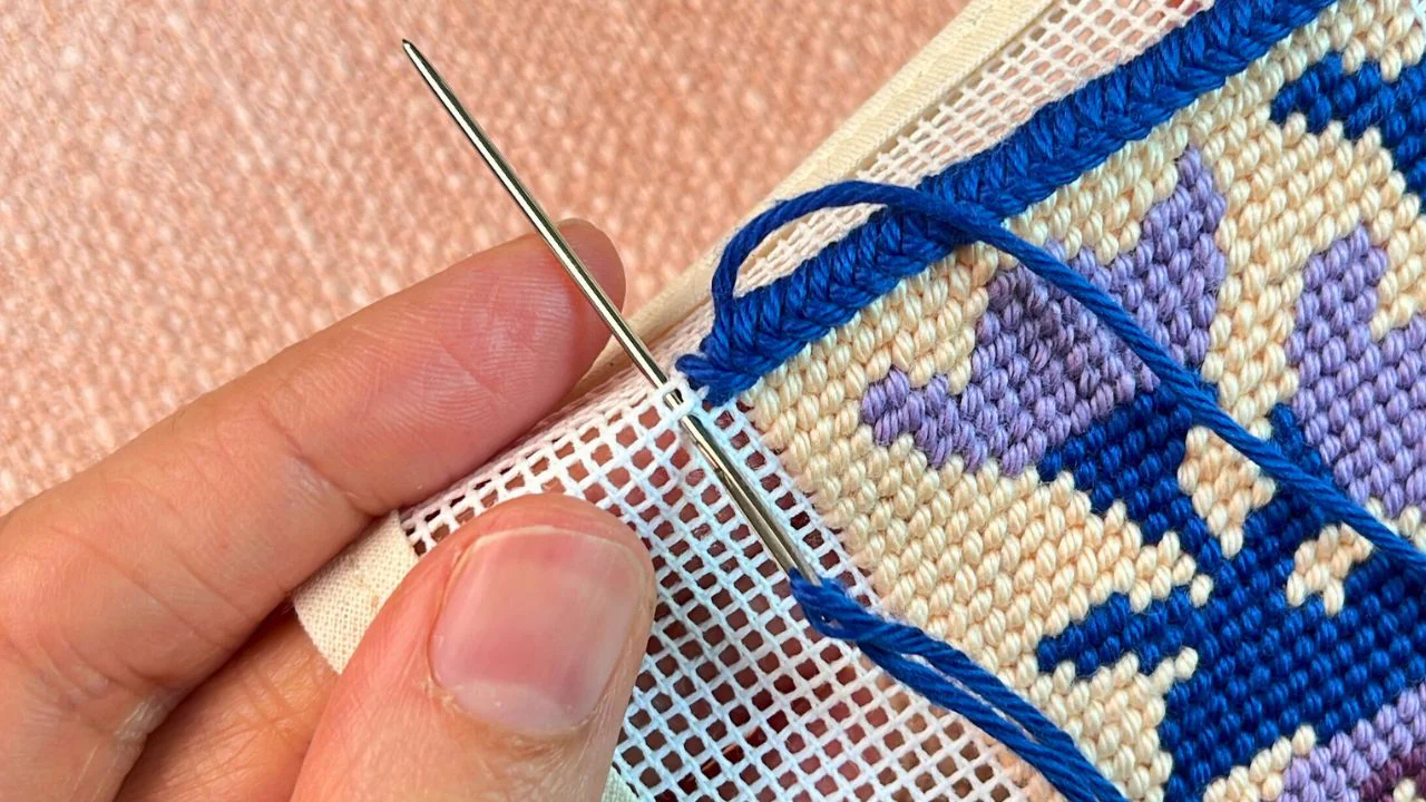 Stitcher holding a binding stitch needlepoint project mid-process with needle in hand