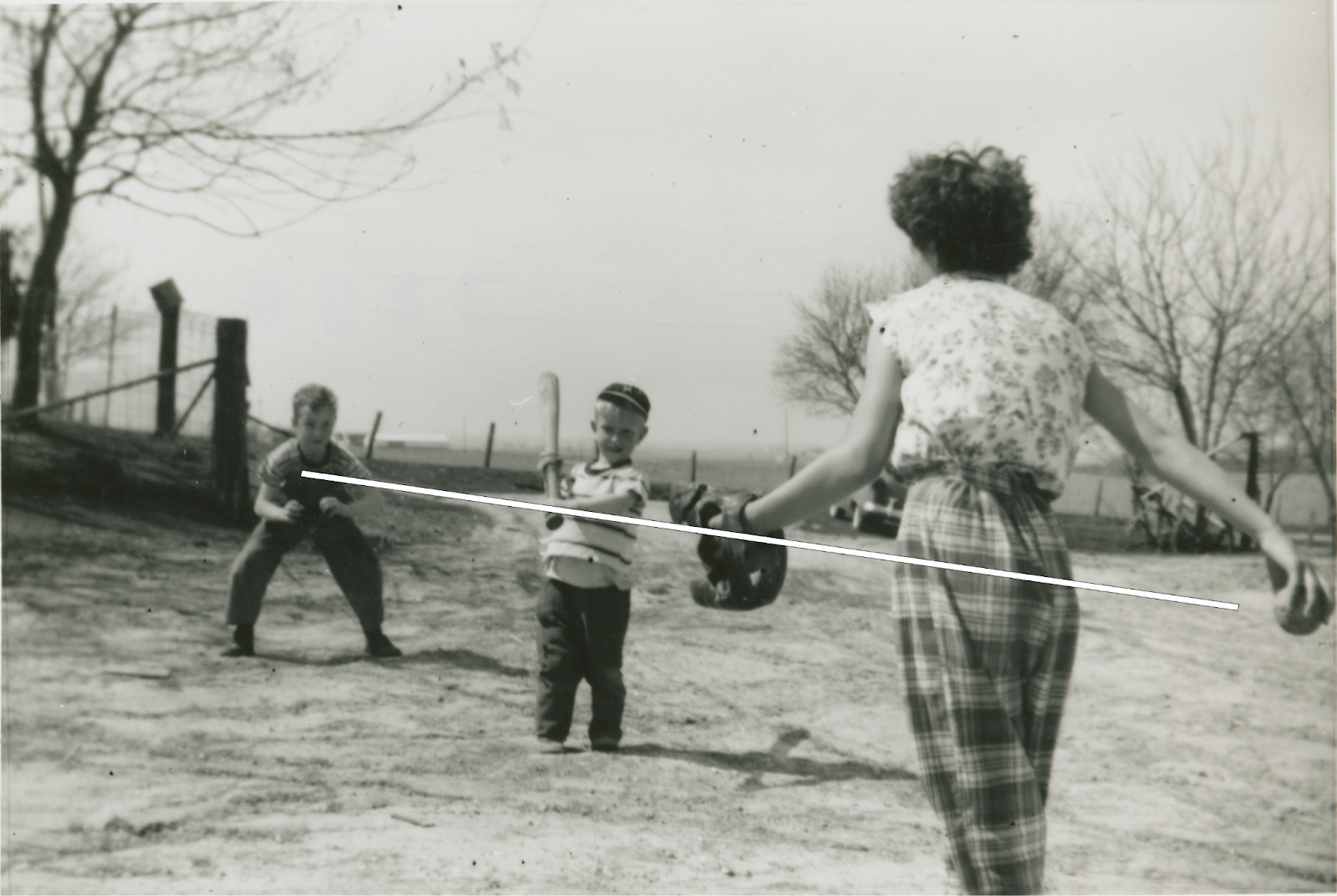 A woman with back toward camera prepares to throw a baseball to a small bow holding a bat, with another boy behind the batter as the catcher