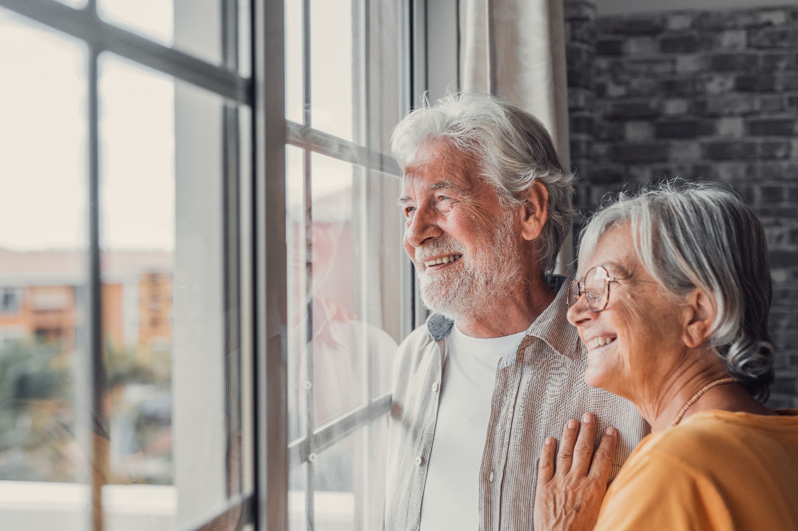 A senior couple smiling while looking out the window of their new home in assisted living.