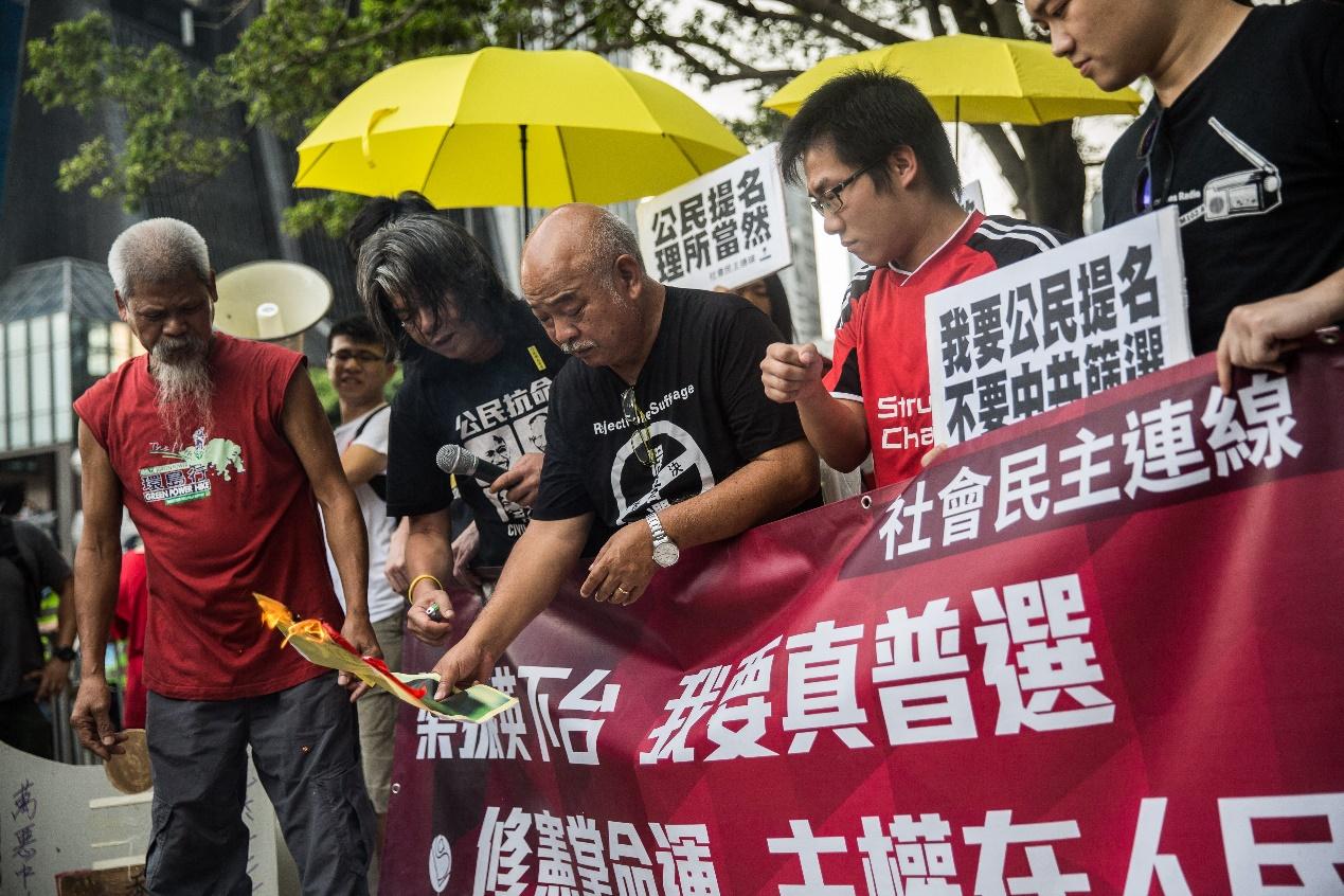 Leung Kwok-hung (2nd L) -- known as "Long Hair" -- of the League of Social Democrats, political activist Koo Sze-yiu (front L) and other activists burn a picture of Hong Kong Chief Executive Leung Chun-ying as they hold a banner calling for him to step down during a rally in Hong Kong on July 1, 2015, on the sidelines of the annual flag raising ceremony to mark the anniversary of Hong Kong's handover from Britain to China. July 1 is traditionally a day of protest in Hong Kong and also marks the anniversary of the handover from Britain to China in 1997, under a "one country, two systems" agreement. AFP PHOTO / ANTHONY WALLACE (Photo by ANTHONY WALLACE / AFP)
