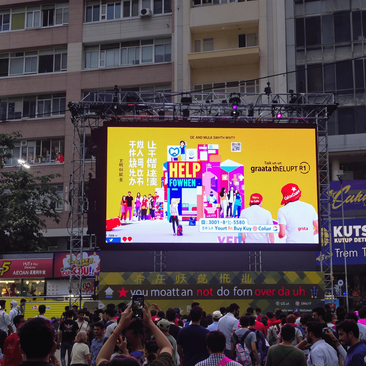 Outdoor event with a large bright LED screen showing colorful ads, surrounded by a lively crowd in an urban setting.