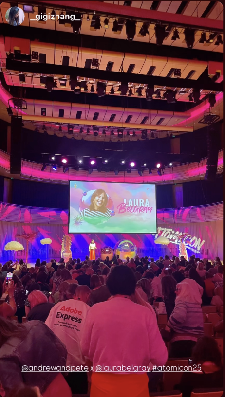 A crowded auditorium is filled with people watching a live presentation at Atomicon, a colorful business and marketing event. The stage is brightly lit with pink, purple, and blue lighting, and a large screen displays an image of Laura Belgray. She stands on stage in front of the screen, speaking to the audience. The decor includes neon signs, palm trees, and bold branding. Text at the bottom credits @andrewandpete, @laurabelgray, and #atomicon25.