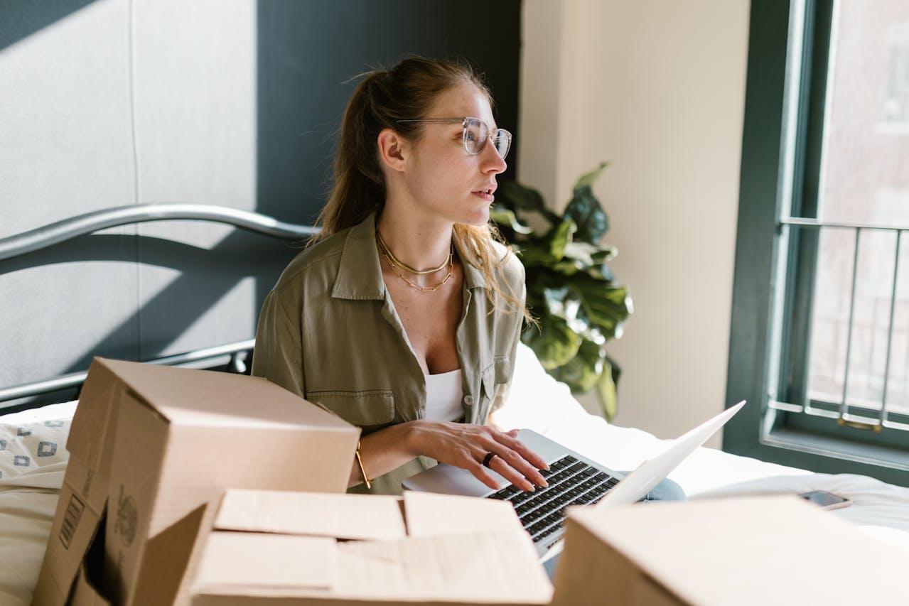 A woman with glasses sitting among the boxes with a laptop in her hands