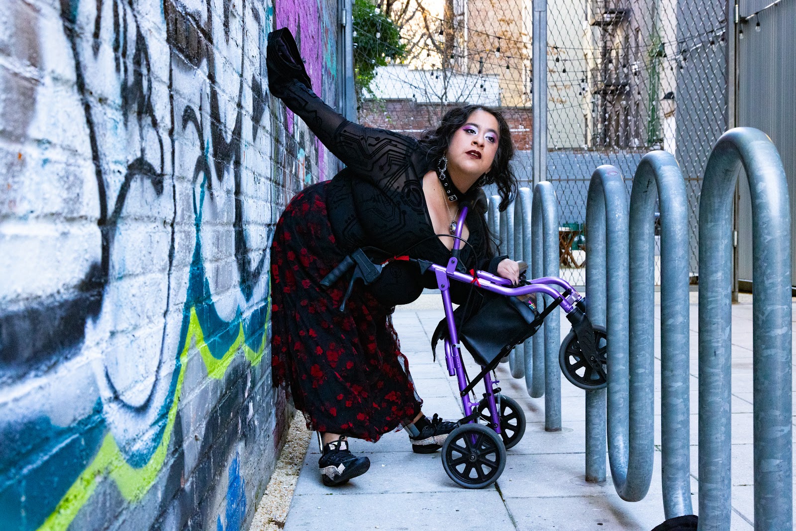 a photo of Vanessa, a chicana woman with caramel skin. She is leaning against a graffiti wall and on top of her tilting walker named Pluto. Her outfit gives dreamy-cyber-gothic-romantic vibes. She is wearing a flowy long sleeve mesh blouse with a digital design. Her black skirt with red flowers is slightly visible. Framing her is a metal looping bike stand
